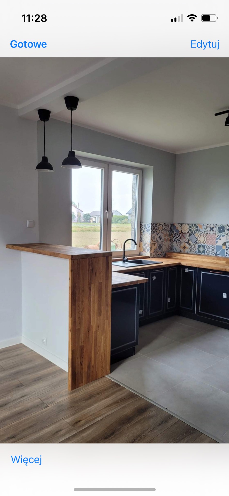 Nowoczesna kuchnia z czarnymi szafkami, drewnianym blatem i barkiem, wzorzystą glazurą nad blatem, widokiem na pole z okna i dwiema czarnymi lampami wiszącymi.