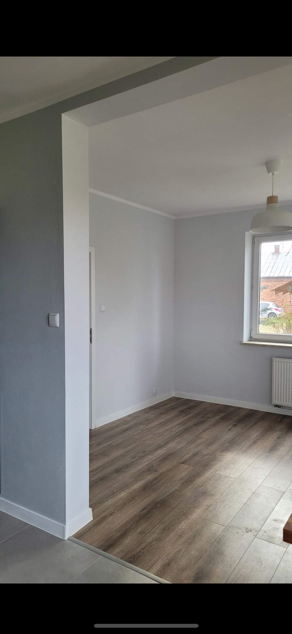 Pomalowane na szaro i biało wnętrze pokoju z panelami podłogowymi, widoczny fragment okna z grzejnikiem i lampą sufitową