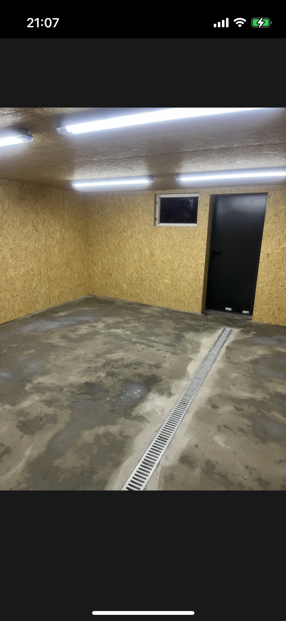 Wnętrze pomieszczenia z surowymi ścianami z płyty OSB, betonową podłogą z odpływem liniowym, oświetleniem LED podsufitowym i czarnymi drzwiami.