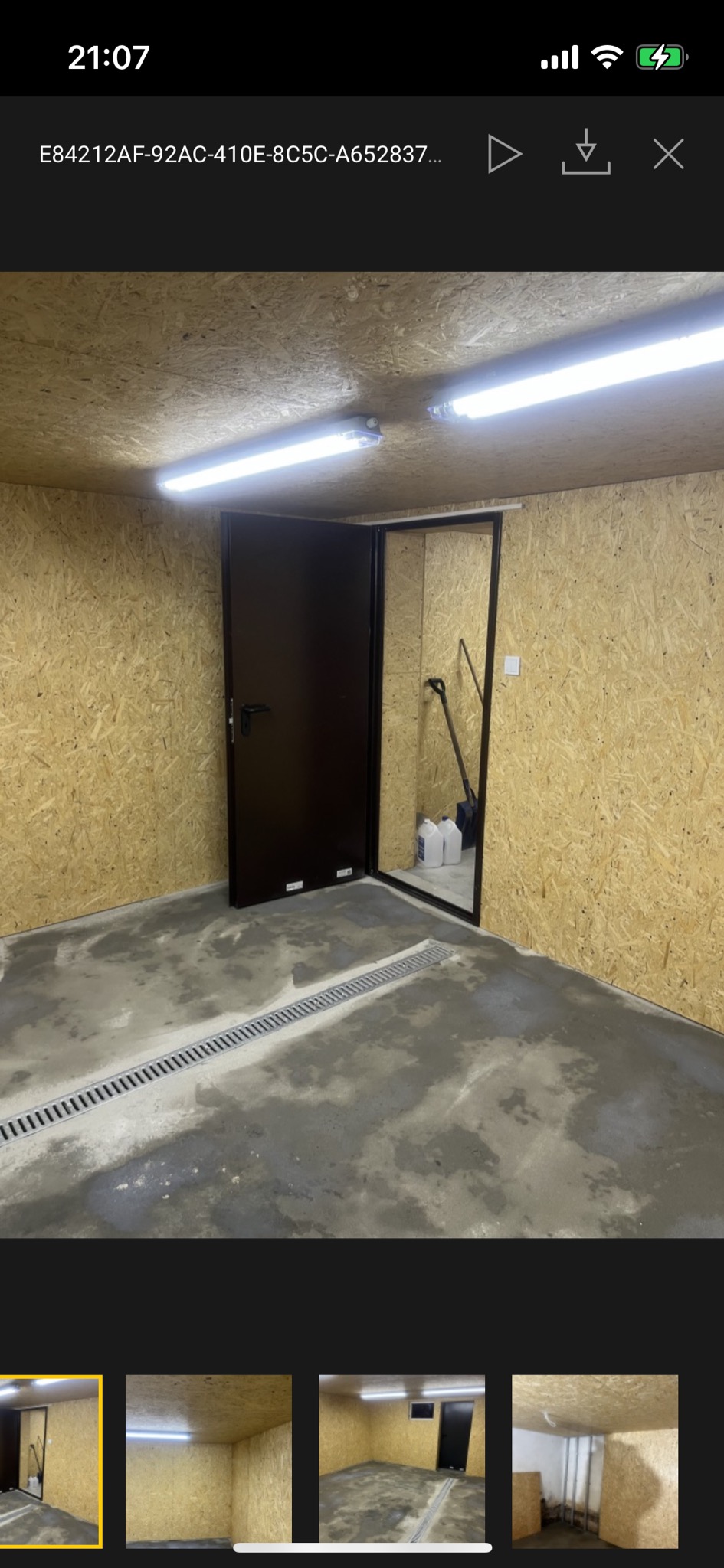 Surowe wnętrze pomieszczenia z ścianami wyłożonymi płytami OSB, betonową podłogą z odpływem liniowym, oświetleniem LED i ciemnymi drzwiami.