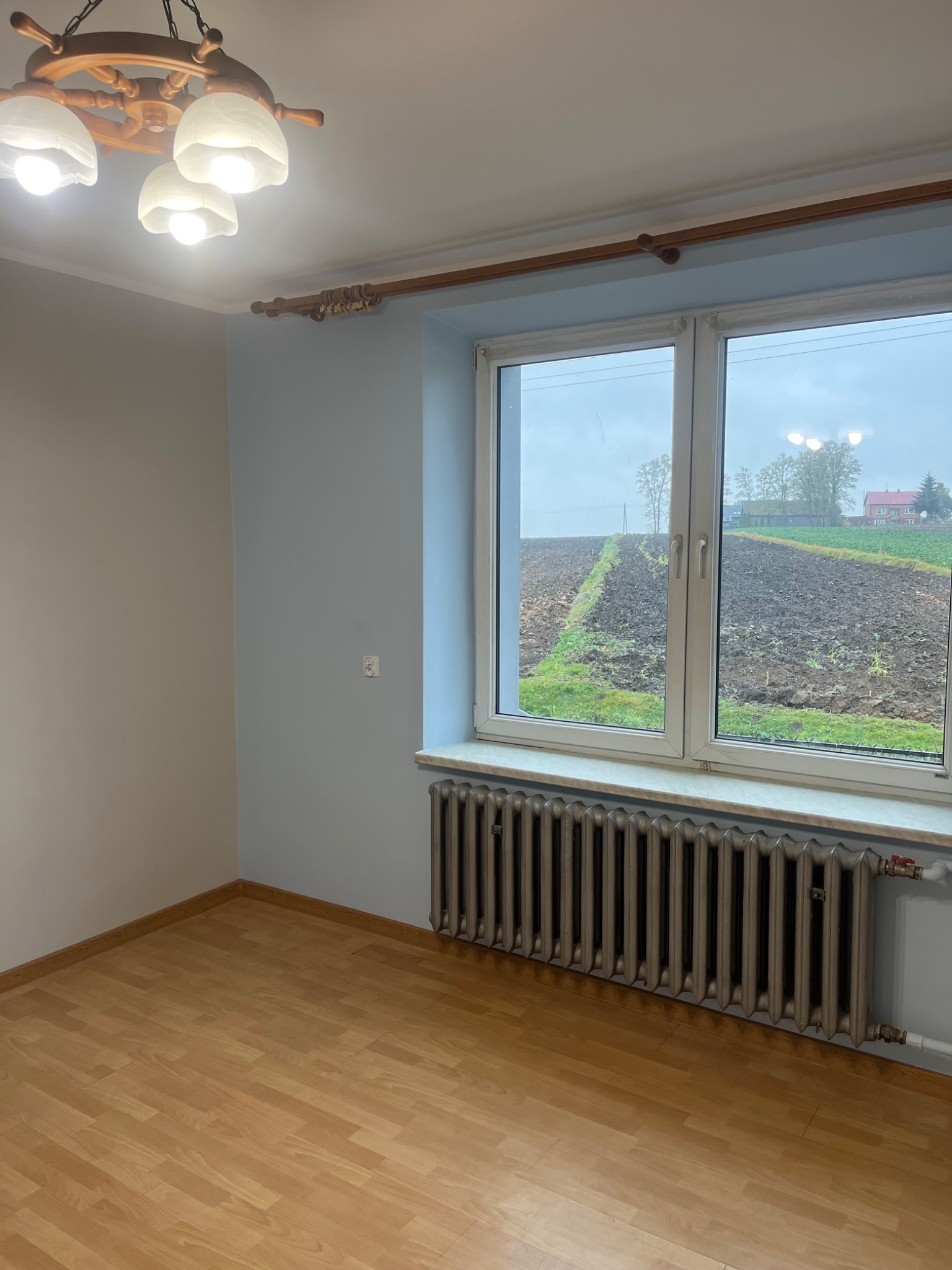 Pusta, jasna przestrzeń z drewnianą podłogą, szarym grzejnikiem pod oknem z widokiem na pole i dom, ściany pomalowane na dwa odcienie szarości, drewniany karnisz i lampa w stylu marynistycznym.