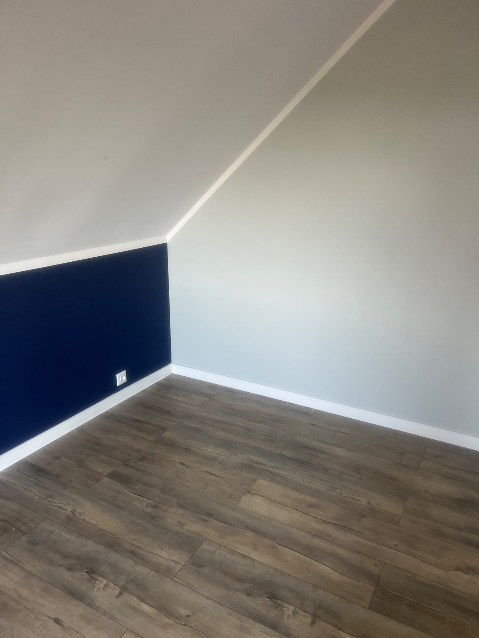 Pomalowane wnętrze poddasza: ściana w kolorze granatowym do połowy wysokości, reszta ściany i sufit w odcieniach szarości, widoczny kontakt elektryczny i panele podłogowe imitujące drewno.