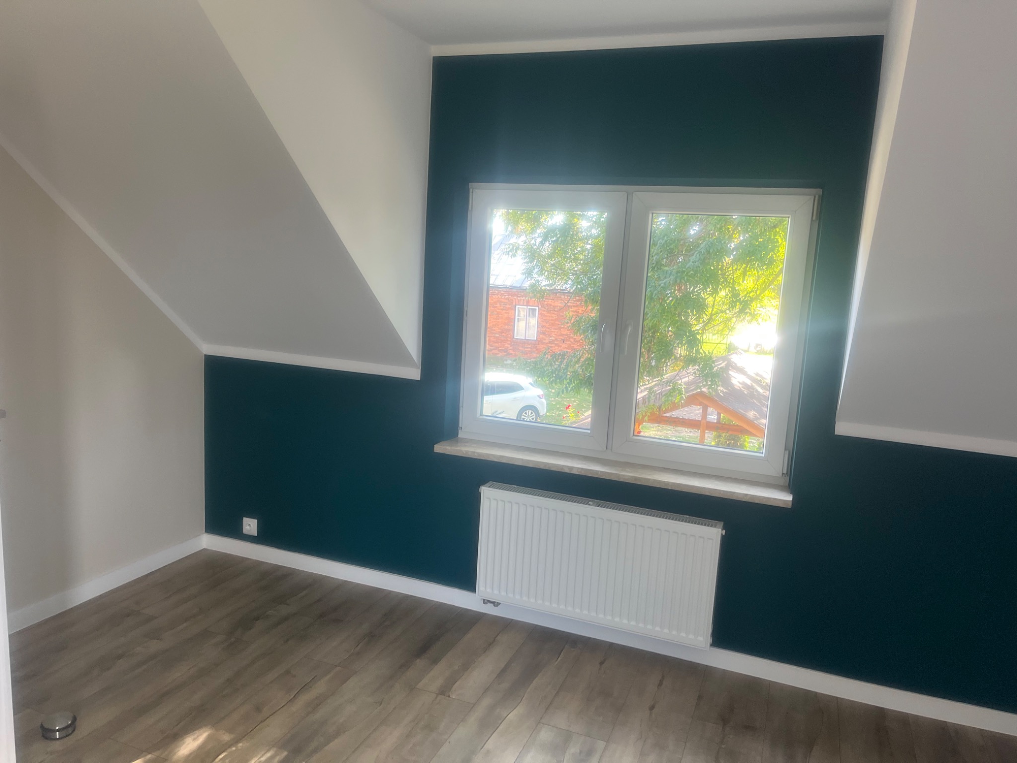 Pomalowane na ciemnozielono ściany w pokoju na poddaszu z oknem, białe, skośne sufity, jasna drewniana podłoga, biały grzejnik pod oknem.