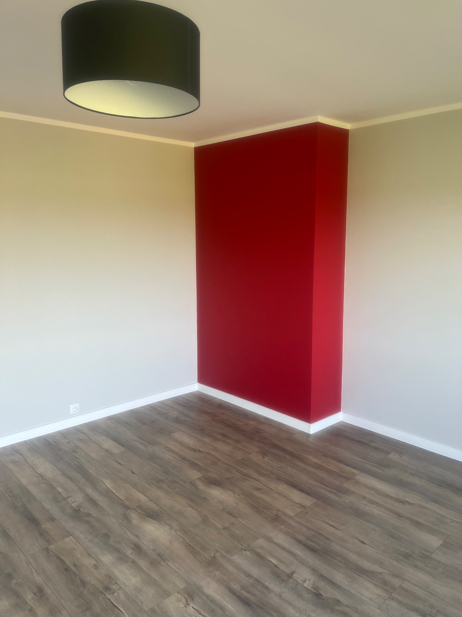 Pomalowany na czerwono narożnik w pokoju z panelami podłogowymi w odcieniach brązu i szarości, widoczny czarny abażur lampy sufitowej.