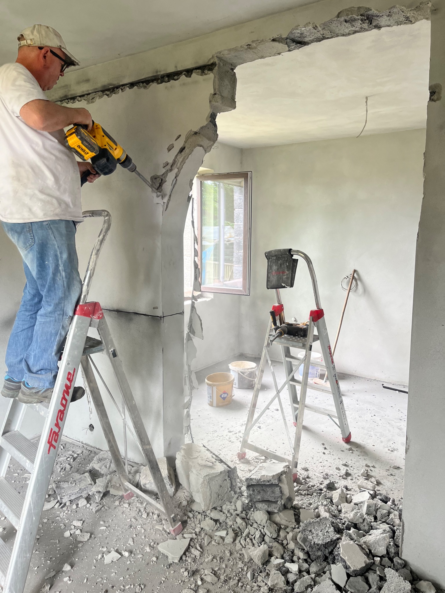 Pracownik na drabinie z żółtym młotem udarowym wykuwa otwór w szarej ścianie, gruz na podłodze, okno w tle.
