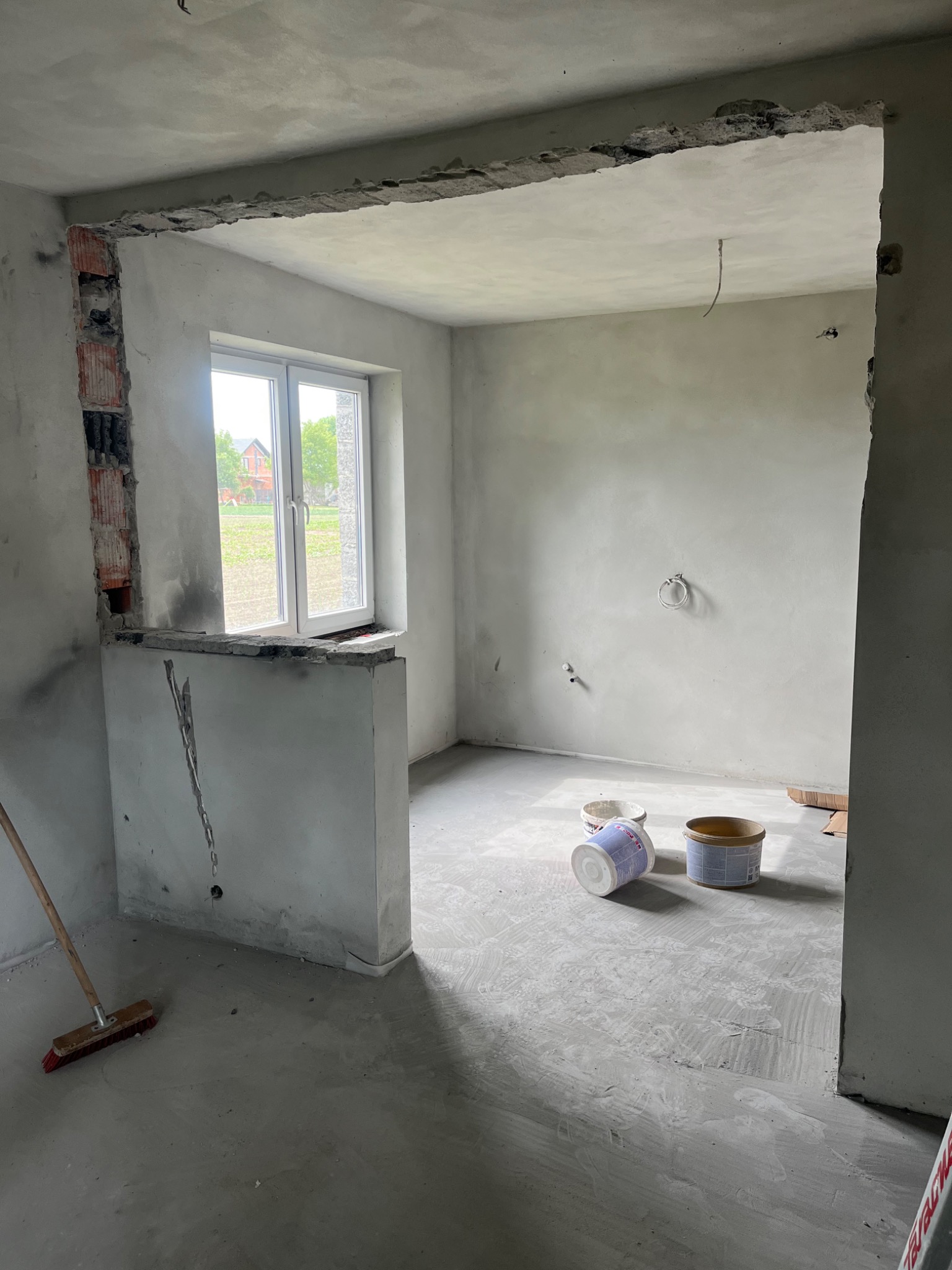 Pokój w trakcie prac wykończeniowych, widoczne ściany po szpachlowaniu, dwa wiadra z farbą na podłodze, widoczne ślady po pracach na podłodze, fragment ściany z odsłoniętą cegłą, okno z widokiem...