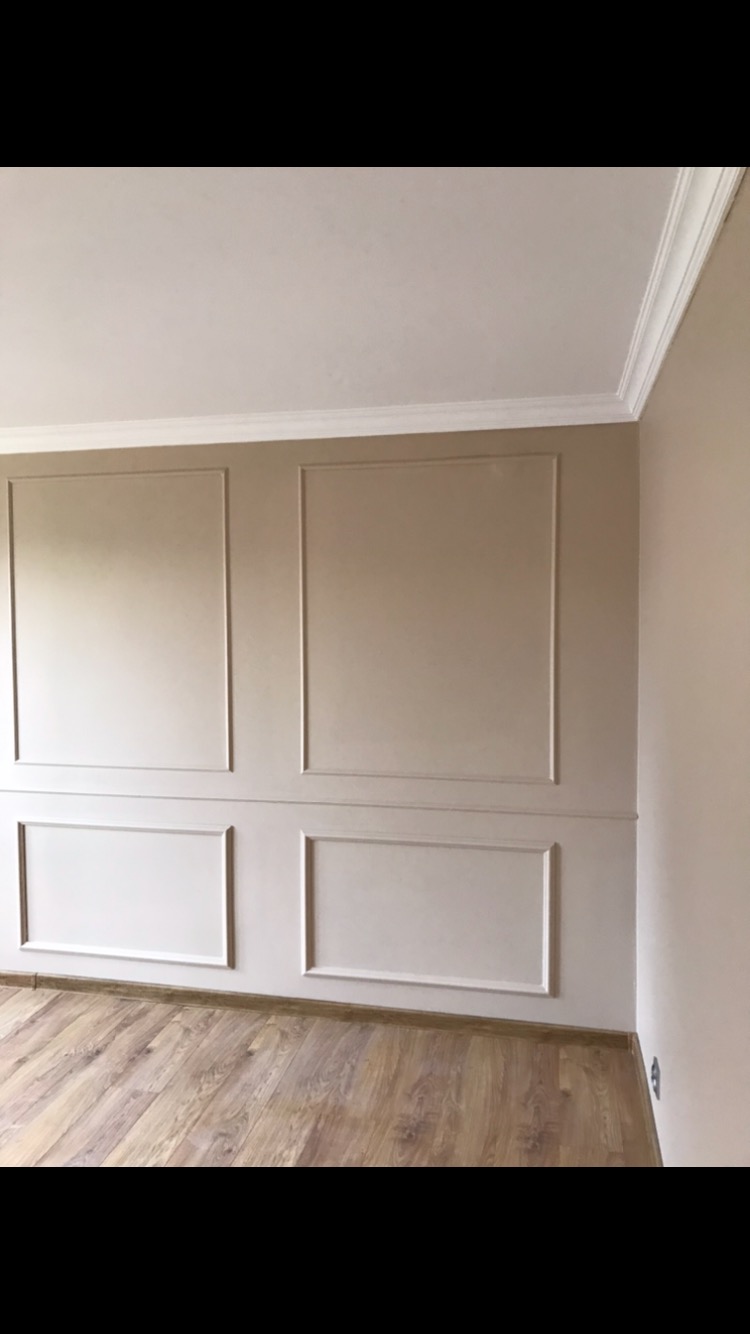 Pomalowane ściany w beżowym odcieniu z dekoracyjnymi ramkami i listwami przypodłogowymi, parkiet na podłodze.