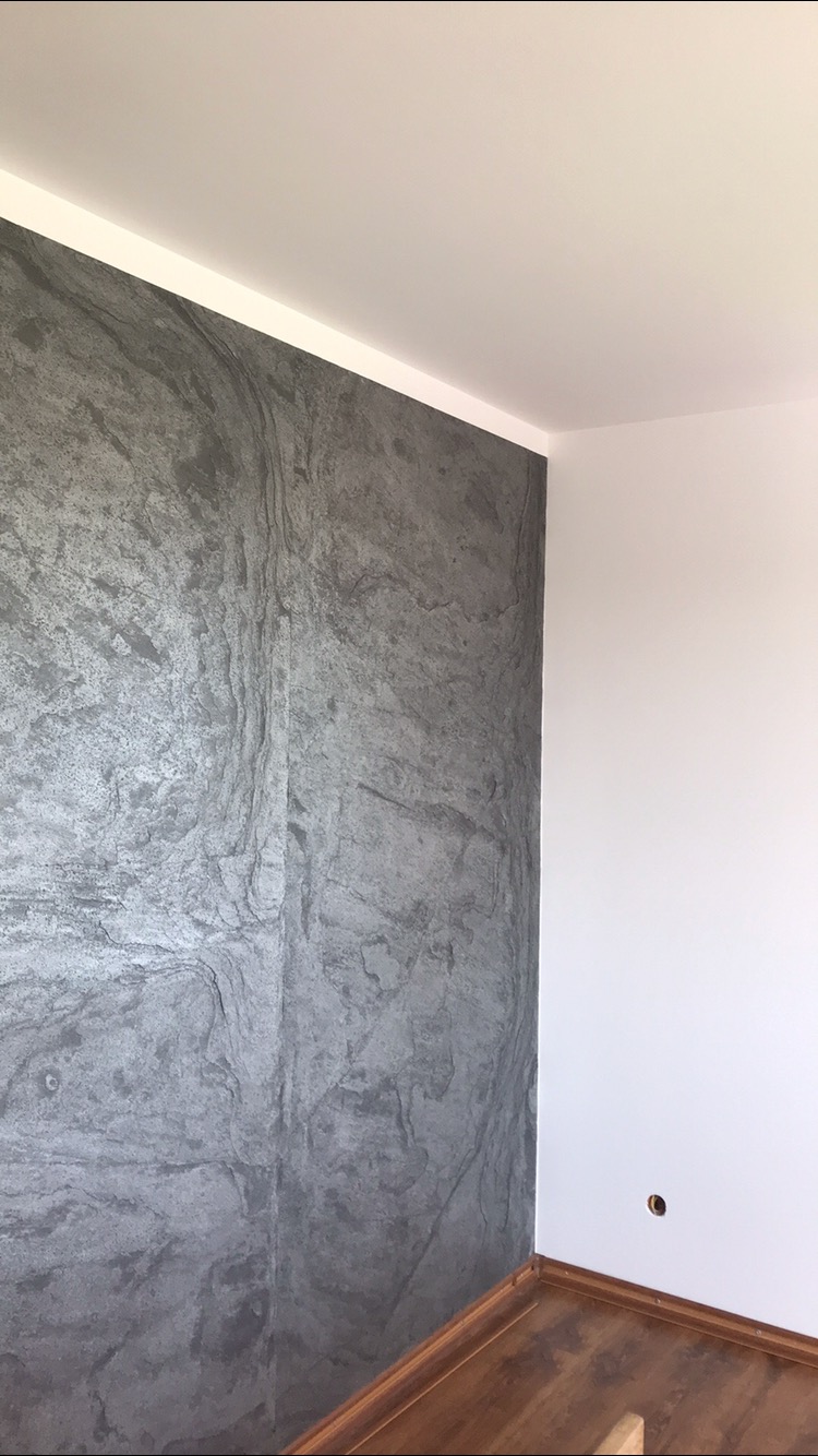 Ściana w salonie wykończona szarą farbą imitującą kamień, kontrastująca z białą ścianą i drewnianą podłogą.