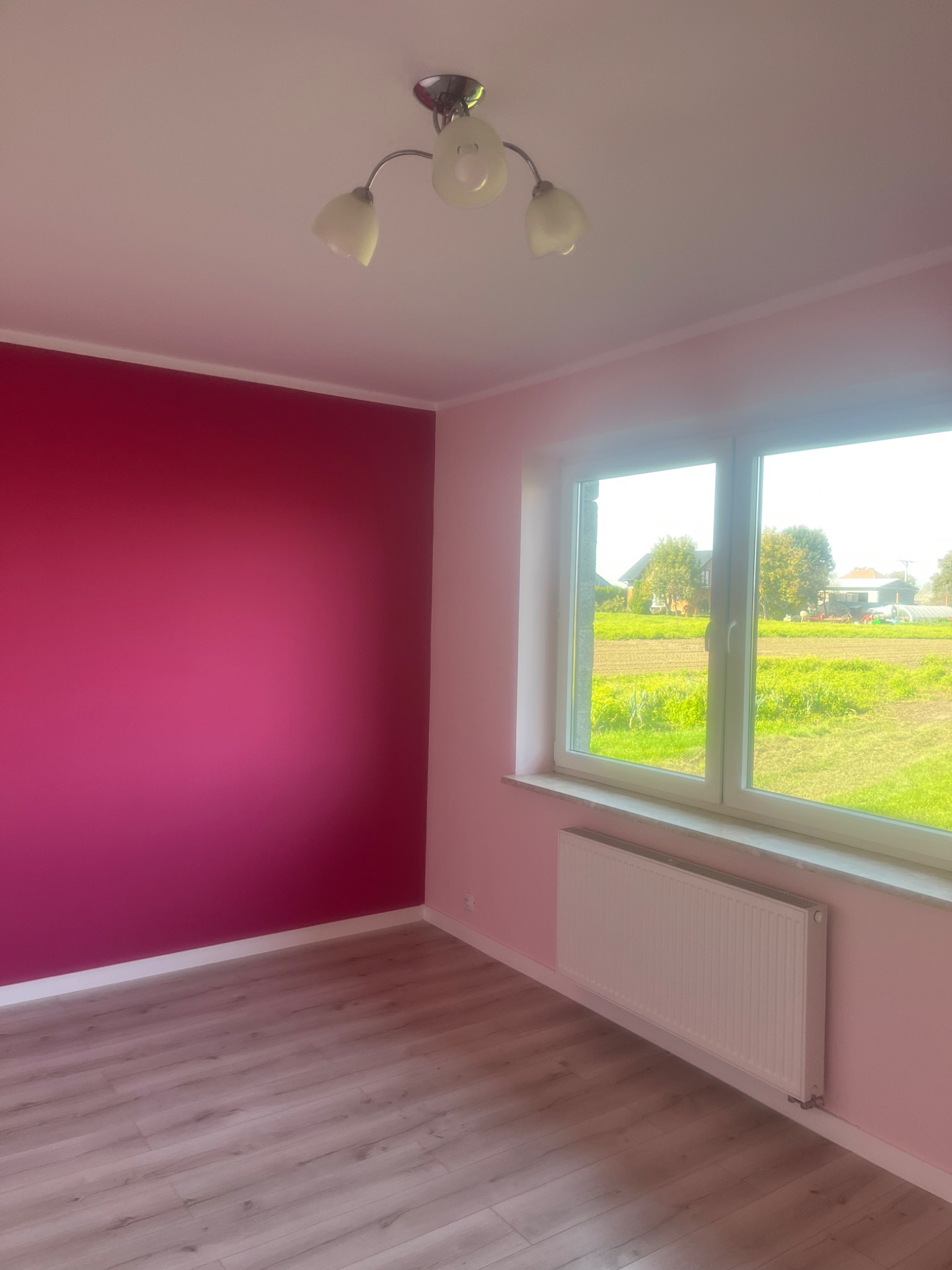 Pusty pokój po odświeżeniu w Proszowicach: ściana pomalowana na intensywny różowy kolor, jasne panele podłogowe, białe okno z widokiem na zielone pole i domy, oraz biały grzejnik pod oknem.