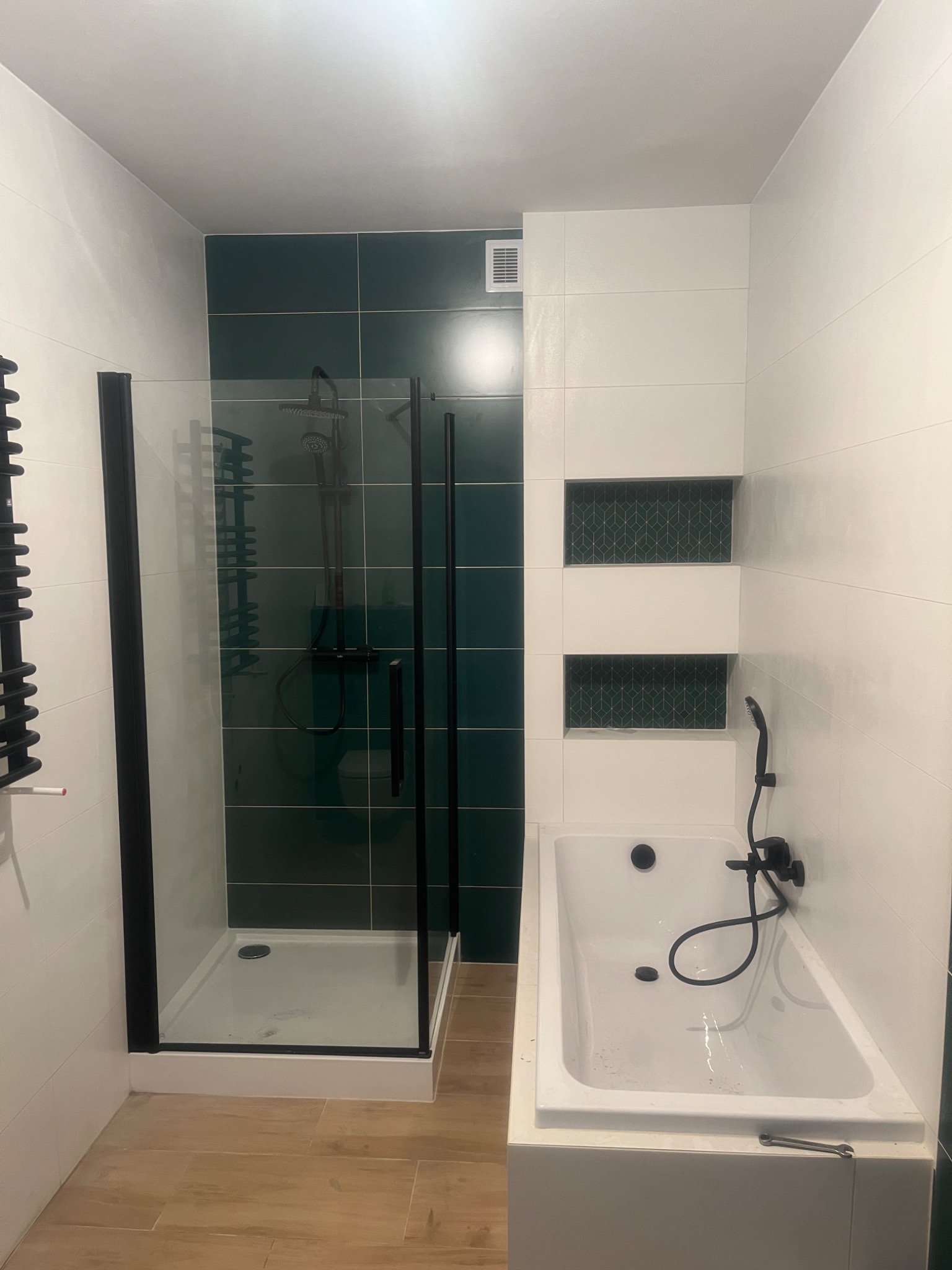 Nowoczesna łazienka z kabiną prysznicową typu walk-in z czarnymi profilami, białą wanną z hydromasażem i ciemnozielonymi płytkami na ścianie prysznica, drewnopodobne płytki podłogowe.