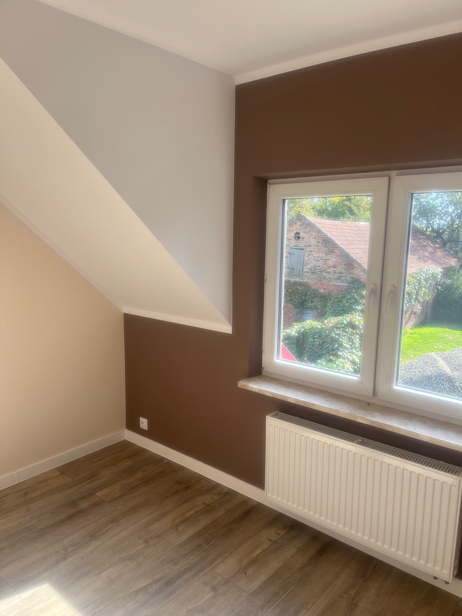Pokój po odświeżeniu: ściany pomalowane na brązowo i beżowo, drewnopodobna podłoga, biały grzejnik pod oknem z widokiem na ceglany budynek porośnięty bluszczem.