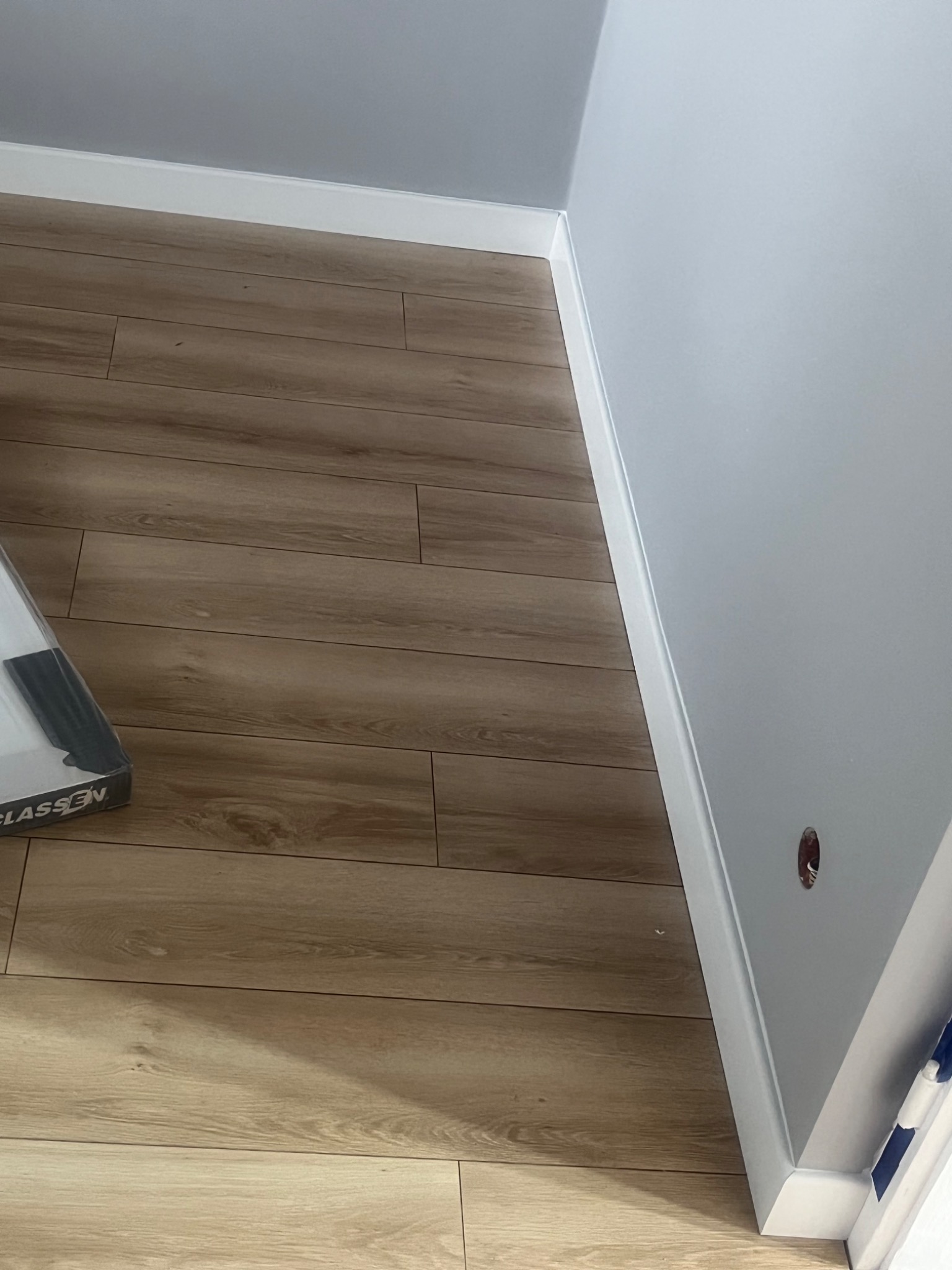 Ukończona podłoga z paneli drewnopodobnych, białe listwy przypodłogowe, świeżo pomalowana szara ściana, widoczny fragment opakowania paneli Classen.