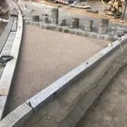 Układanie kostki granitowej w kształcie trójkąta z widocznymi krawężnikami granitowymi i podsypką piaskową. W tle ułożone kostki brukowe i maszyny budowlane.