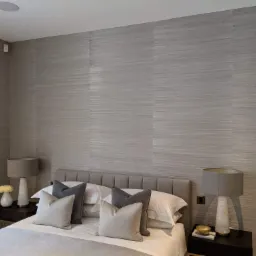 Sypialnia z łóżkiem i szafkami nocnymi, ściany pokryte szarą tapetą o subtelnym, poziomym wzorze przypominającym tkaninę, okrągły głośnik sufitowy w rogu.