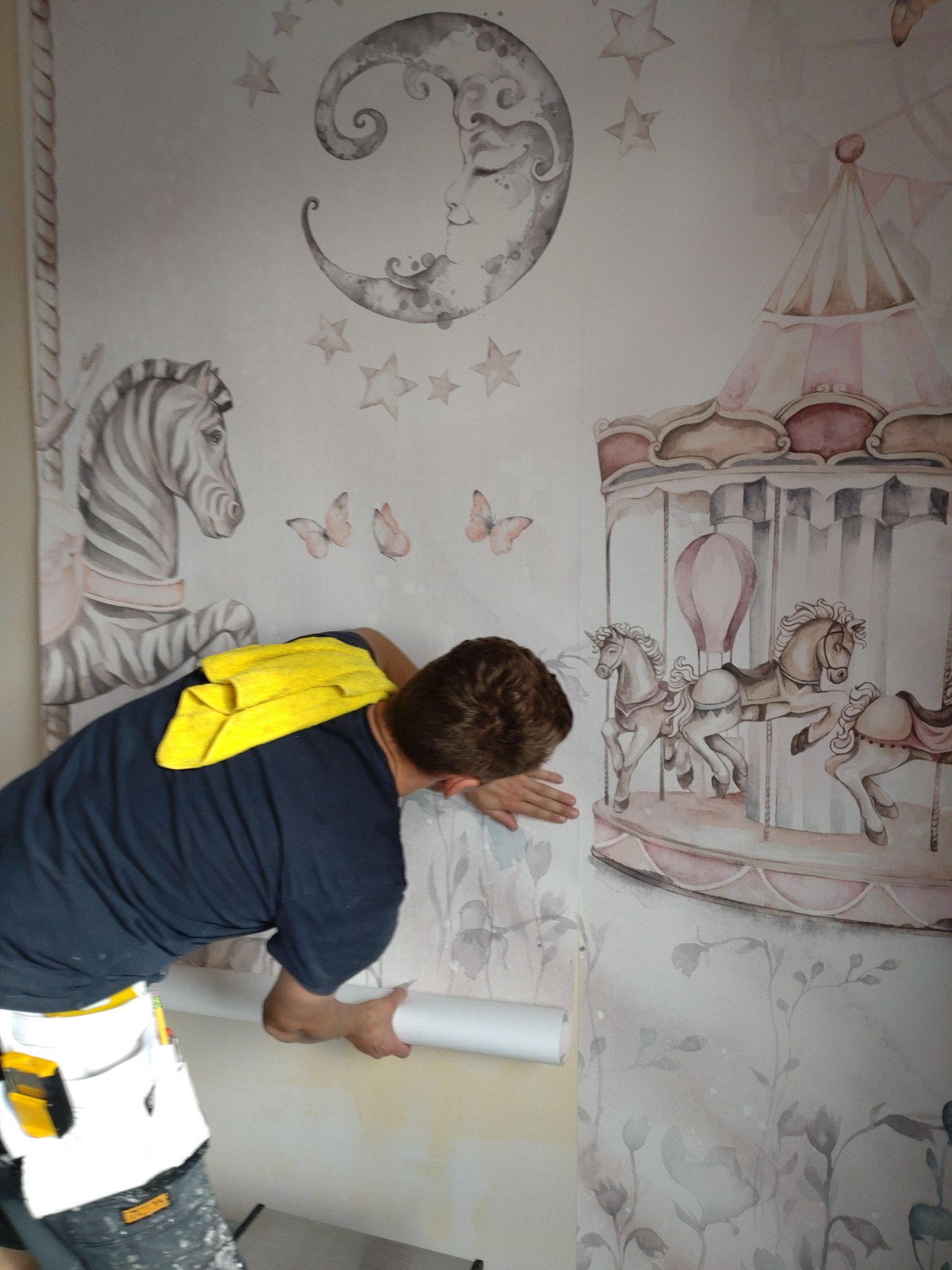 Mężczyzna w trakcie nakładania tapety z motywem karuzeli i księżyca na ścianę, wygładza fragment żółtą ściereczką, widoczny pas tapety w rolce i narzędzia w pasie.