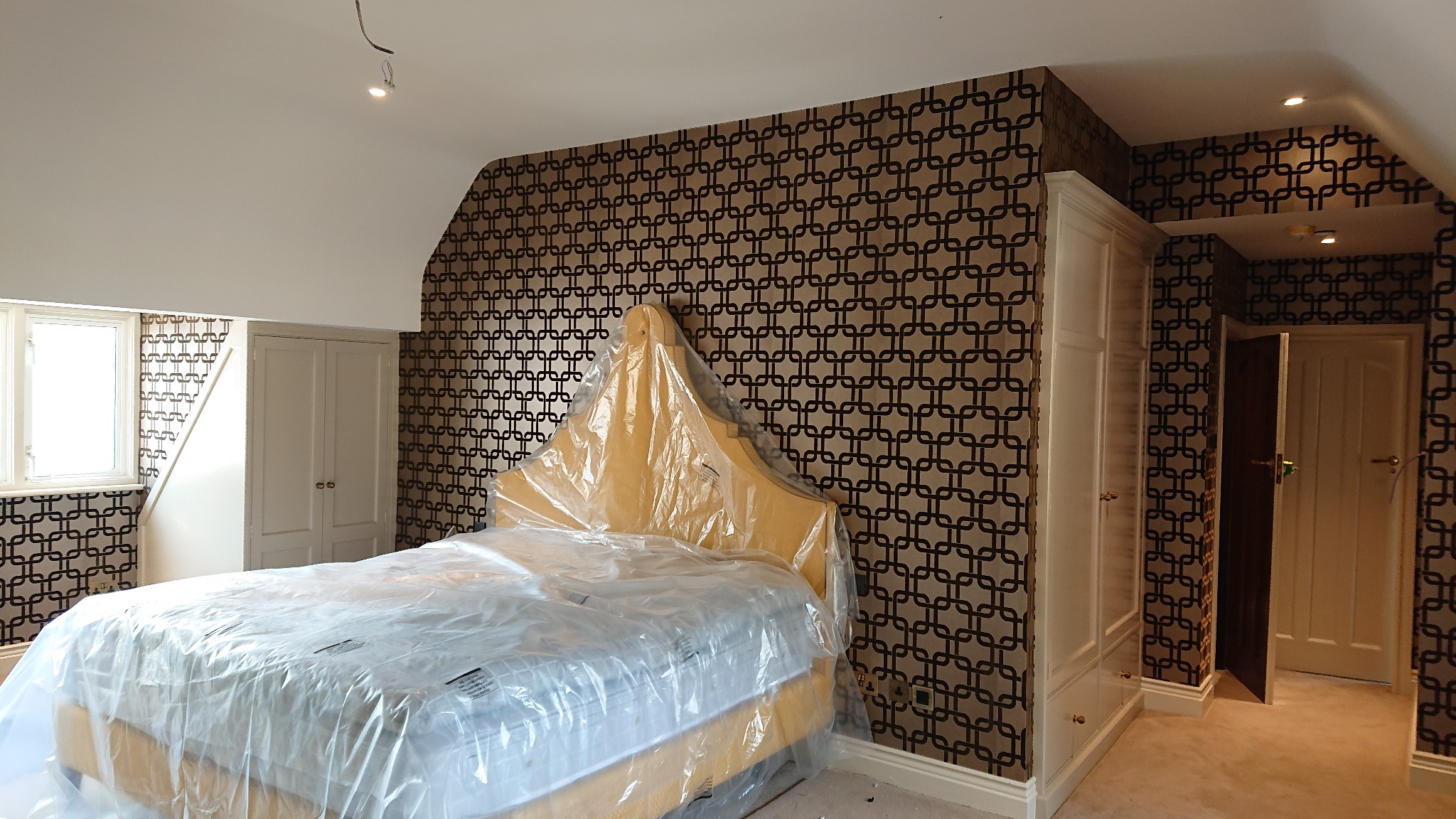 Pokój przed zdjęciem folii ochronnej z łóżka po tapetowaniu ścian wzorzystą tapetą w odcieniach brązu i beżu, widoczne wnękowe szafy w kolorze kremowym.