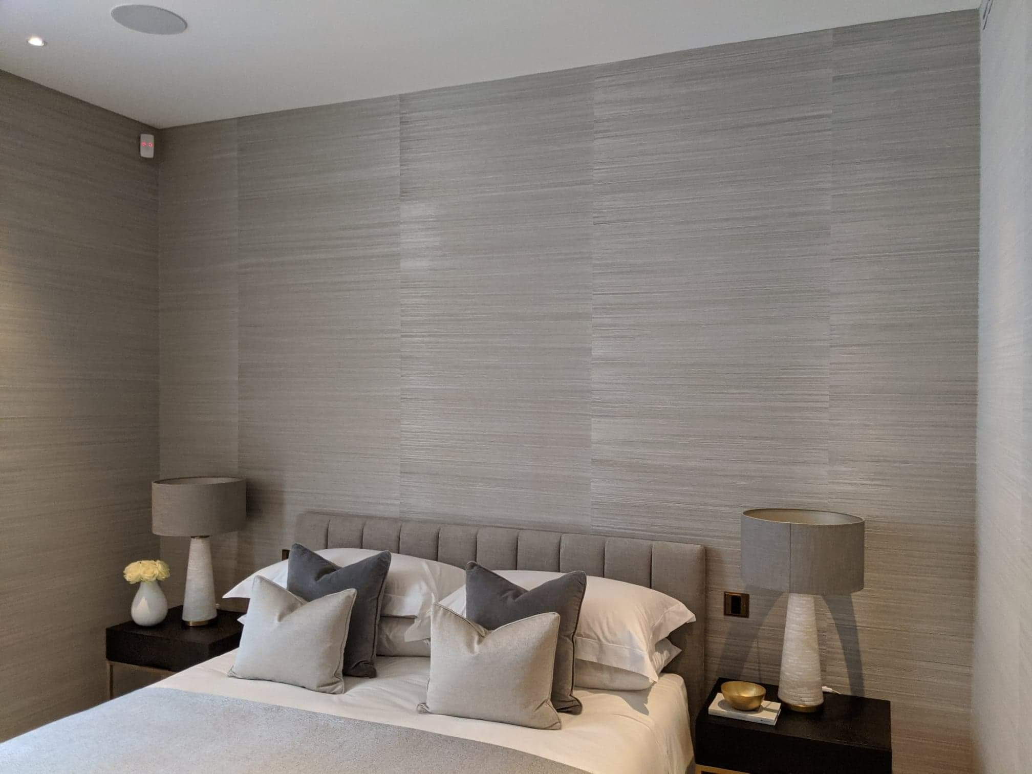 Sypialnia z łóżkiem i szafkami nocnymi, ściany pokryte szarą tapetą o subtelnym, poziomym wzorze przypominającym tkaninę, okrągły głośnik sufitowy w rogu.