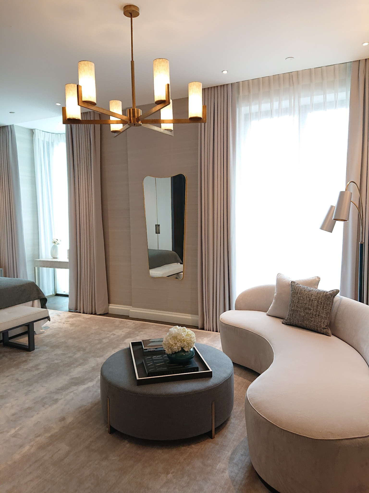 Eleganckie wnętrze pokoju dziennego w odcieniach beżu i szarości z zaokrągloną sofą, okrągłym pufem pełniącym rolę stolika kawowego, nowoczesną lampą podłogową i lustrem w złotej ramie.