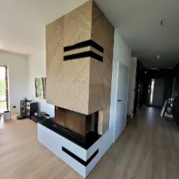 Nowoczesny kominek z obudową z drewnianych paneli ułożonych w geometryczny wzór, z czarnymi wstawkami wentylacyjnymi, umieszczony w jasnym salonie z drewnianą podłogą.