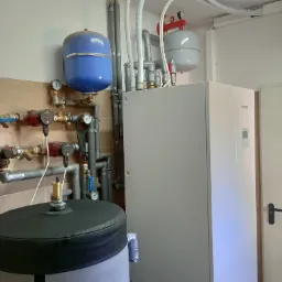 Pompa ciepła Kaisai z osprzętem, w tym zbiornik akumulacyjny w szarej izolacji, niebieski i szary zbiornik wyrównawczy, orurowanie i manometry na ścianie wyłożonej płytkami.