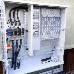 Otwarta, biała skrzynka elektryczna z zainstalowanymi bezpiecznikami, kablami i akcesoriami montażowymi, zamontowana na ścianie budynku. Widoczne przewody z numeracją.