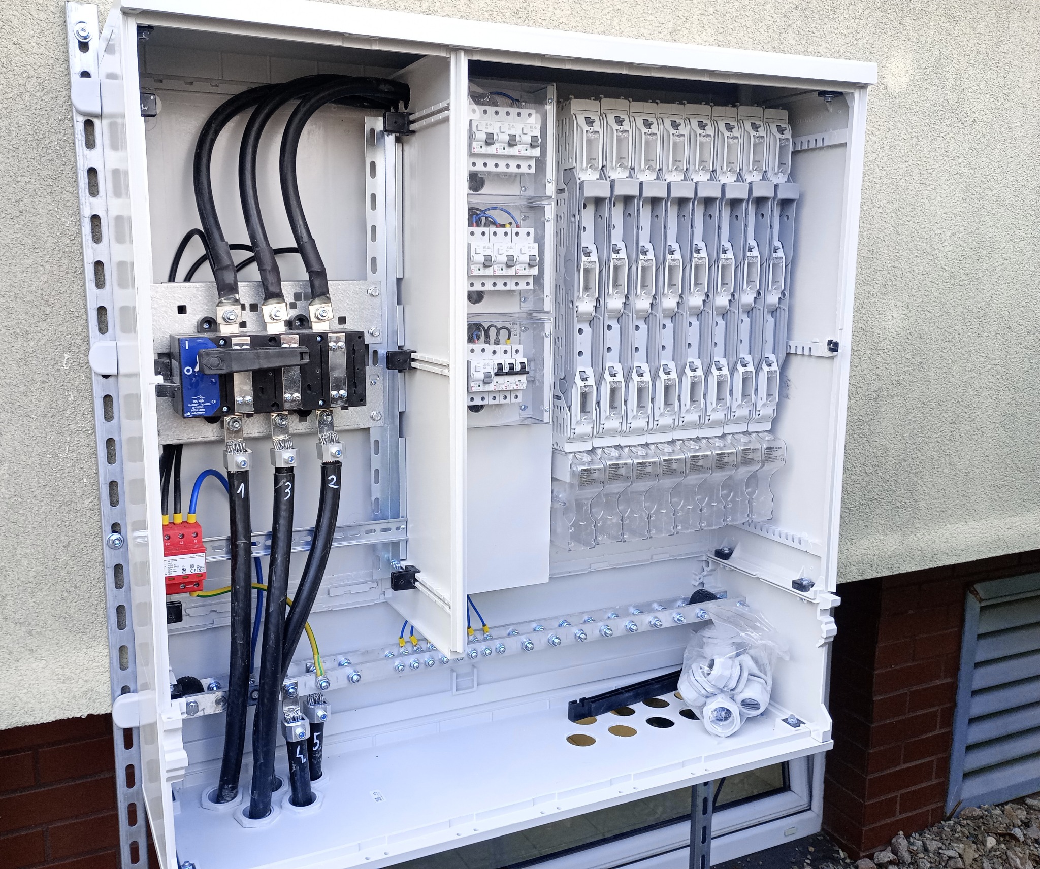 Otwarta, biała skrzynka elektryczna z zainstalowanymi bezpiecznikami, kablami i akcesoriami montażowymi, zamontowana na ścianie budynku. Widoczne przewody z numeracją.