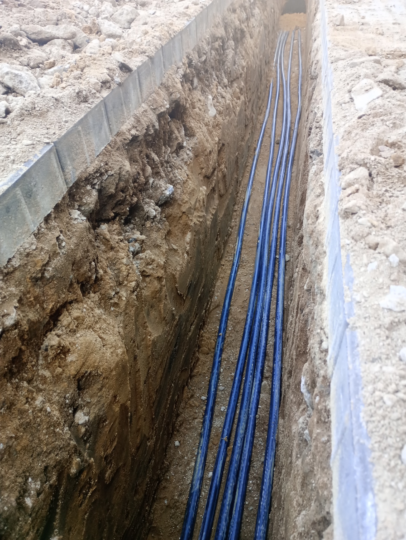 Wykop z ułożonymi niebieskimi rurami kablowymi, widok z góry. Rury równolegle ułożone w wąskim wykopie ziemnym, zabezpieczonym obrzeżami betonowymi.