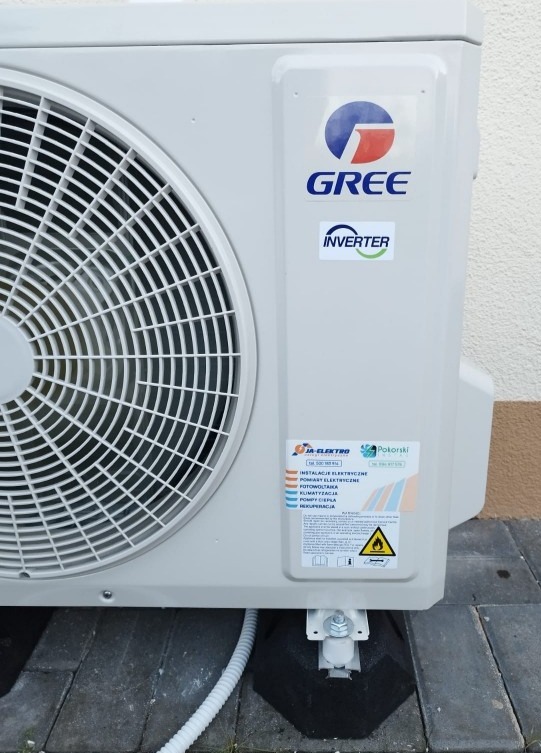 Zewnętrzna jednostka klimatyzacji marki Gree z widocznym logo i naklejką 'Inverter', zamontowana na czarnych podstawkach na kostce brukowej, z widoczną tabliczką informacyjną firmy instalacyjnej...