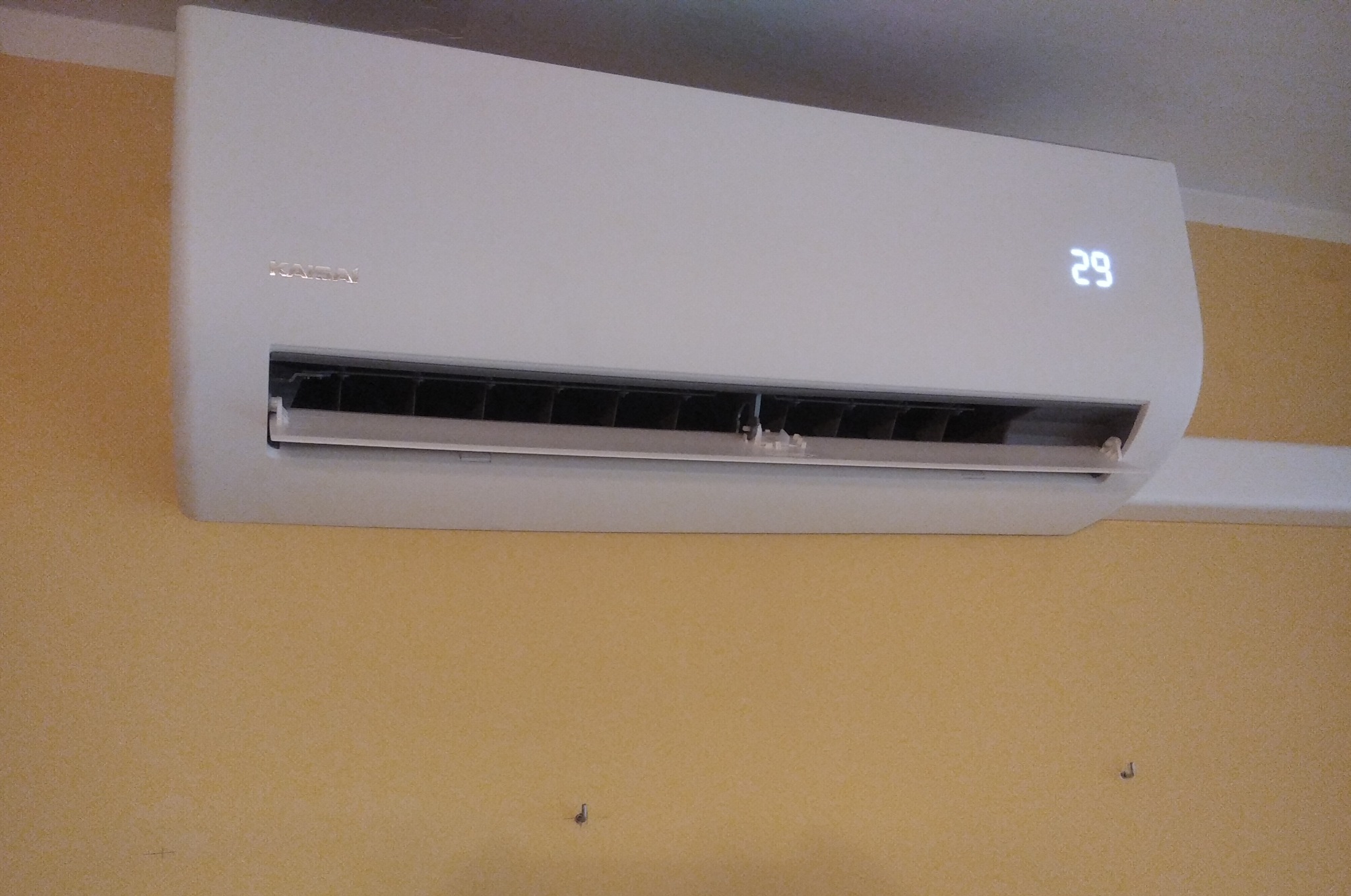 Biała jednostka klimatyzacji marki KAISAI zamontowana na żółtej ścianie, wyświetlająca temperaturę 29 stopni Celsjusza.