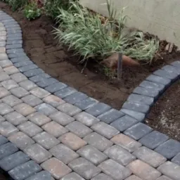 Kwadratowa kostka w odcieniach szarości i brązu ułożona w łukowaty chodnik przylegający do rabaty z trawami ozdobnymi, obrzeże z ciemnoszarej kostki.