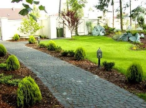 Kręta ścieżka z ciemnej kostki prowadząca przez zadbany ogród z trawnikiem, ozdobnymi krzewami i elementami skalnymi w tle.