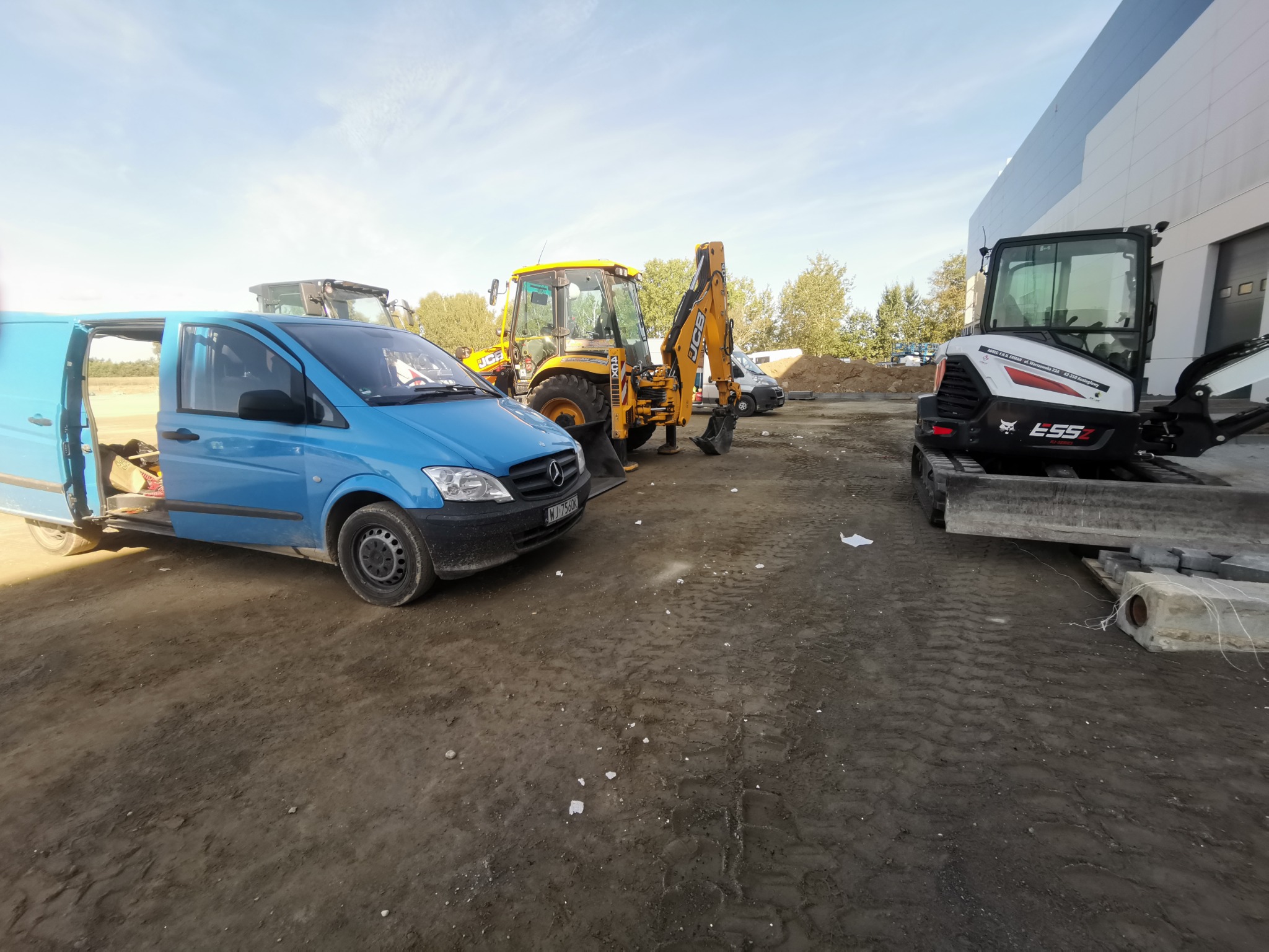 Niebieski van Mercedes z otwartymi drzwiami, żółta koparko-ładowarka JCB i biała mini-koparka Bobcat ESSZ na placu budowy, widoczne ślady opon i porozrzucane materiały.