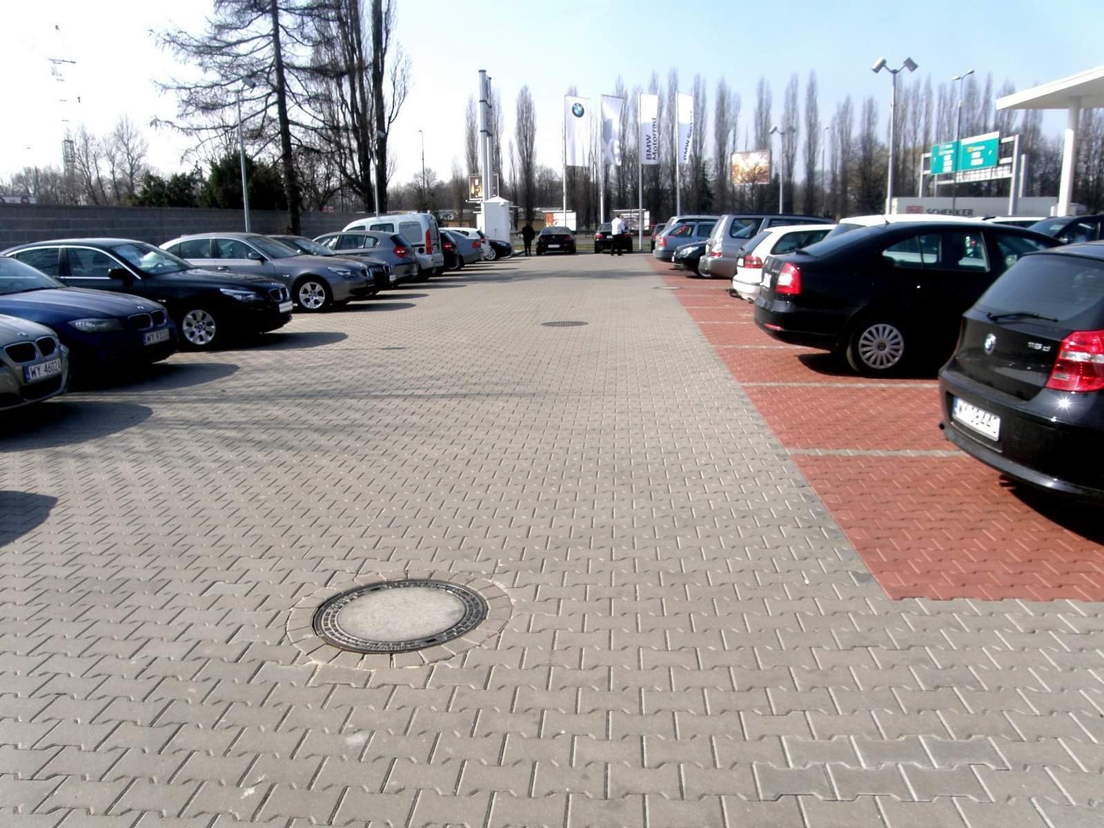 Szary parking wyłożony kostką brukową typu 'I', z rzędem zaparkowanych samochodów i studzienką kanalizacyjną na pierwszym planie.