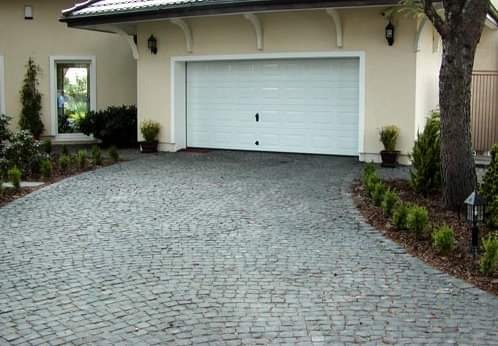 Podjazd do garażu wyłożony szarą kostką granitową ułożoną w łukowaty wzór, z widocznym fragmentem domu, drzewem i niskimi krzewami po bokach.