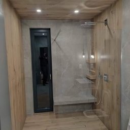 MASTERHOUSE TRL SP.Z.O. O - Nowoczesna kabina prysznicowa typu walk-in z drewnopodobnymi płytkami na ścianach i podłodze, szklana ścianka, wbudowane półki i siedzisko, minimalistyczny design.