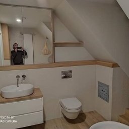 MASTERHOUSE TRL SP.Z.O. O - Nowoczesna łazienka na poddaszu ze skośnym sufitem, białą umywalką nablatową na szafce z drewnianym blatem, podwieszaną toaletą i lustrem w szafce, w której odbija się osoba robiąca zdjęcie.