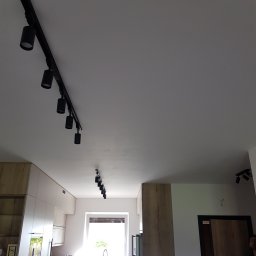 MASTERHOUSE TRL SP.Z.O. O - Nowoczesne oświetlenie szynowe z czarnymi reflektorami w jasnym wnętrzu, widoczne szafki kuchenne z drewnianymi frontami i białe ściany.