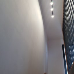 MASTERHOUSE TRL SP.Z.O. O - Świeżo pomalowana ściana w jasnym odcieniu szarości, oświetlona trzema punktowymi lampami sufitowymi, z widoczną częścią nowoczesnej balustrady schodowej z pionowych, ciemnych elementów.