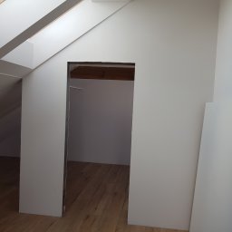 MASTERHOUSE TRL SP.Z.O. O - Poddasze po malowaniu ścian na biało, widoczne drewniane belki stropowe oraz podłoga z paneli, surowe obramowanie otworu drzwiowego.