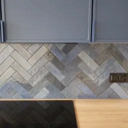 Błękitno-szara mozaika ułożona w jodełkę na ścianie nad blatem kuchennym, z widocznym gniazdkiem elektrycznym i fragmentem płyty indukcyjnej.