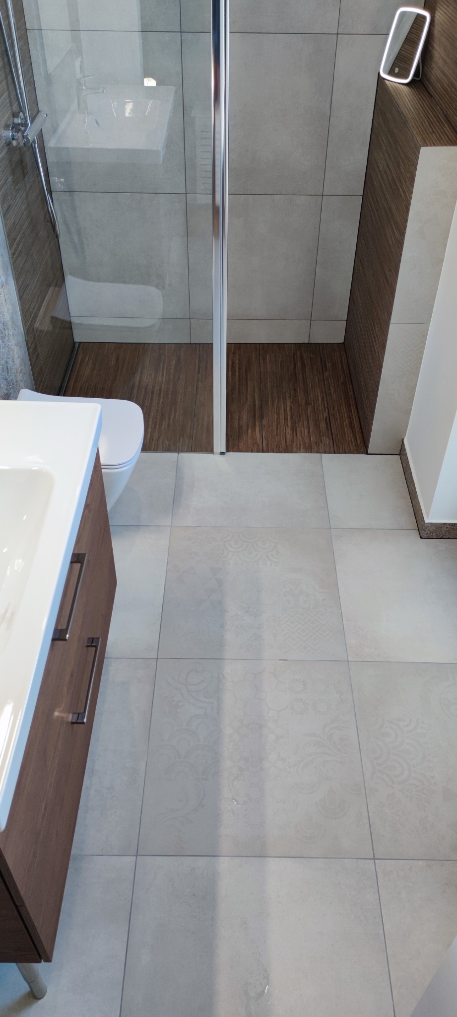 Nowoczesna łazienka po remoncie z kabiną prysznicową, drewnopodobnymi płytkami w strefie natrysku, umywalką z szafką w odcieniach brązu i jasnymi płytkami podłogowymi z delikatnym wzorem.