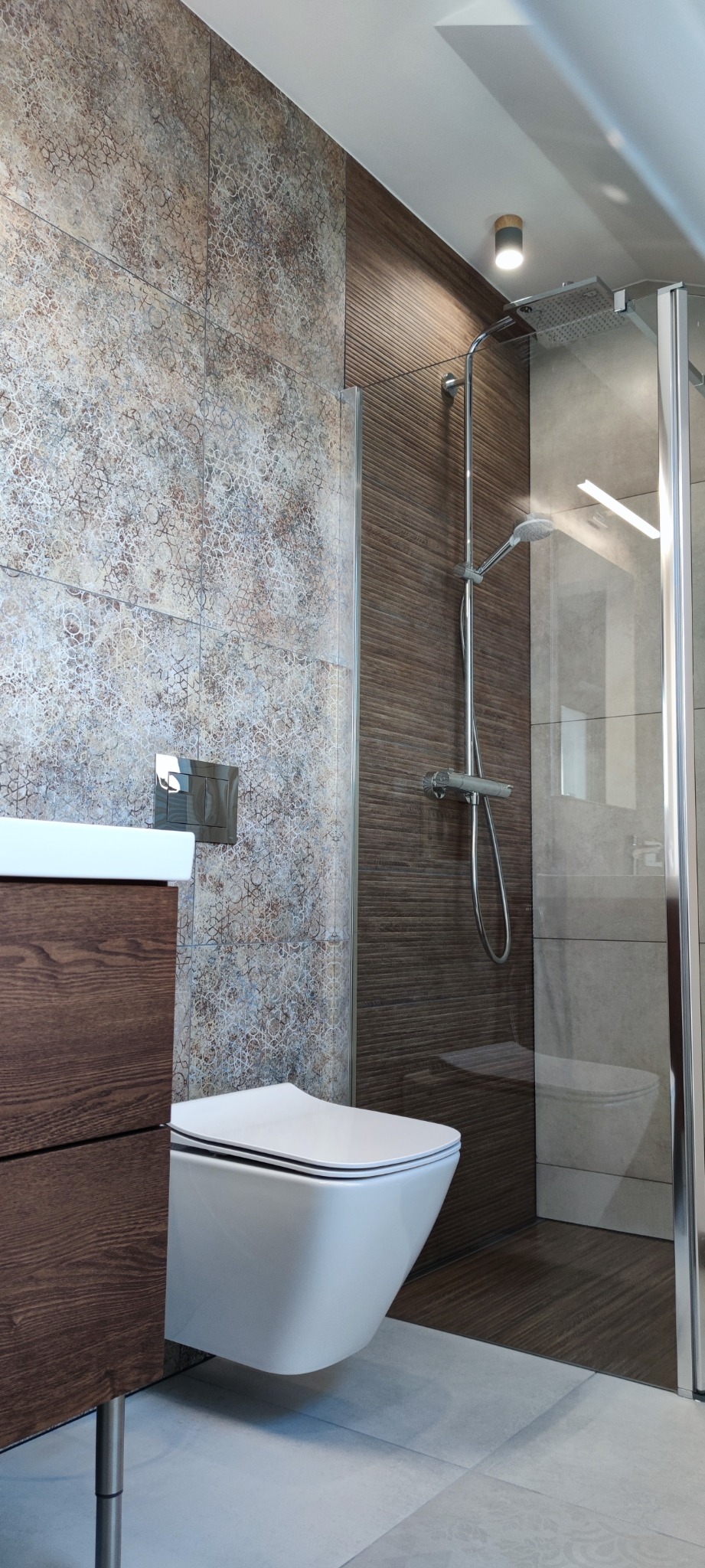 Nowoczesna łazienka z wiszącą miską WC, kabiną prysznicową typu walk-in i płytkami w odcieniach brązu i beżu z delikatnym wzorem.