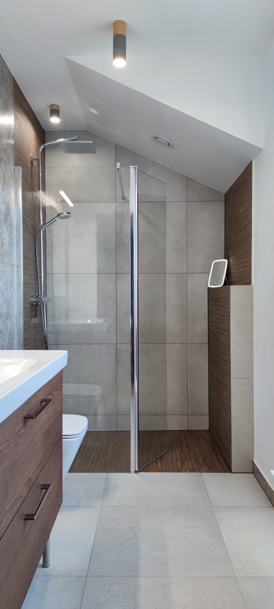 Nowoczesna łazienka z kabiną prysznicową typu walk-in, szarą okładziną ścienną imitującą beton i drewnianymi akcentami, oświetlona punktowymi lampami sufitowymi.