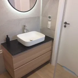 Nowoczesna łazienka z umywalką nablatową, czarnym blatem i drewnianą szafką z szufladami, okrągłe lustro z podświetleniem LED, minimalistyczny design, szara płytka imitująca kamień na ścianach.