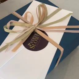 Niebieskie pudełko z białym kartonem, przewiązane wstążką w odcieniach beżu i brązu, z okrągłą naklejką z logo 'Patryk Słowi Fotografia', obok nożyczki.