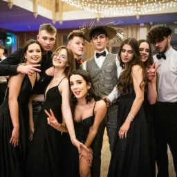 Grupa elegancko ubranych młodych ludzi pozuje do zdjęcia na tle sali balowej. Wśród nich osoba w słomkowym kapeluszu i muszce. Parkiet i dekoracje świetlne w tle.