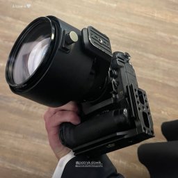 Czarny aparat z dużym obiektywem trzymany w dłoni, widoczny fragment białej koszuli i czarnego garnituru. Ujęcie z bliska, na jasnym tle.