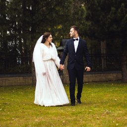 Patryk Słowik Fotografia - Para młoda w Aleksandrowie spaceruje po trawie, trzymając się za ręce. Panna młoda w białej sukni i futerku, pan młody w granatowym garniturze z muchą. Romantyczny kadr.