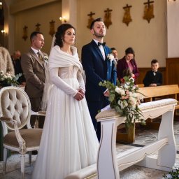 Patryk Słowik Fotografia - Para młoda w kościele podczas ceremonii ślubnej. Panna młoda w białej sukni, pan młody w granatowym garniturze. W tle goście i elementy wystroju kościoła.