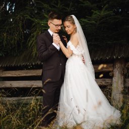 Patryk Słowik Fotografia - Młoda para w eleganckich strojach ślubnych, on w okularach, ona w sukni z welonem, czule trzymająca się za ręce na tle drewnianego płotu i iglaków, wśród wysokiej trawy.